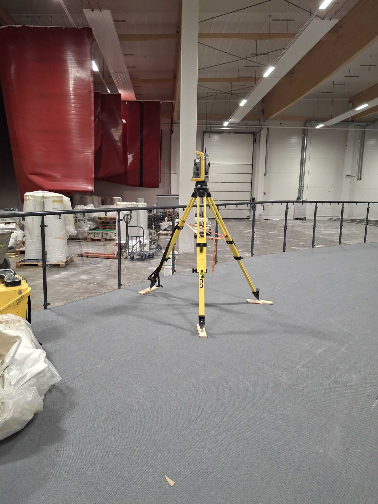 Keltainen mittausjalusta mittauslaitteineen harmaalle sisälattialle varastossa, jossa on varastolavoja ja punaisia ​​riippuvia verhoja.